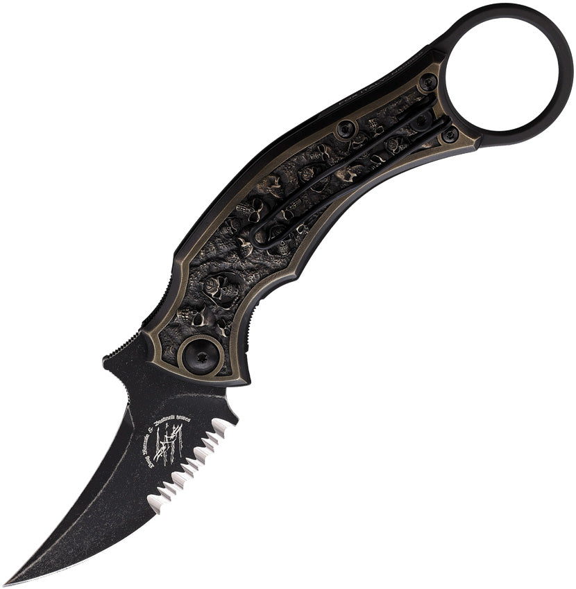 A folding knife with a black stonewash finish partially serrated blade and a sculpted bronze handle featuring skull relief artwork. The knife includes a finger ring and a pocket clip.