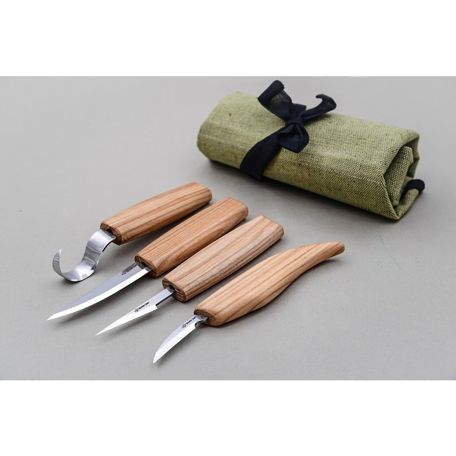 A set of four wood carving knives with wooden handles, accompanied by a rolled-up canvas with a dark green interior and a black bow on top. The knives are positioned in front of the rolled canvas. Beaver Craft | King of Knives e-store