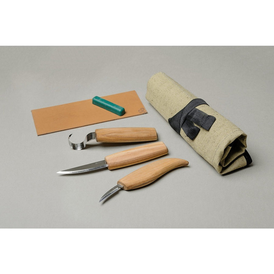 A set of wood carving tools including a sloyd knife, chip carving knife, hook knife, and a leather strop with polishing compound, displayed next to a rolled-up canvas tool roll. Beavercraft | King of knives